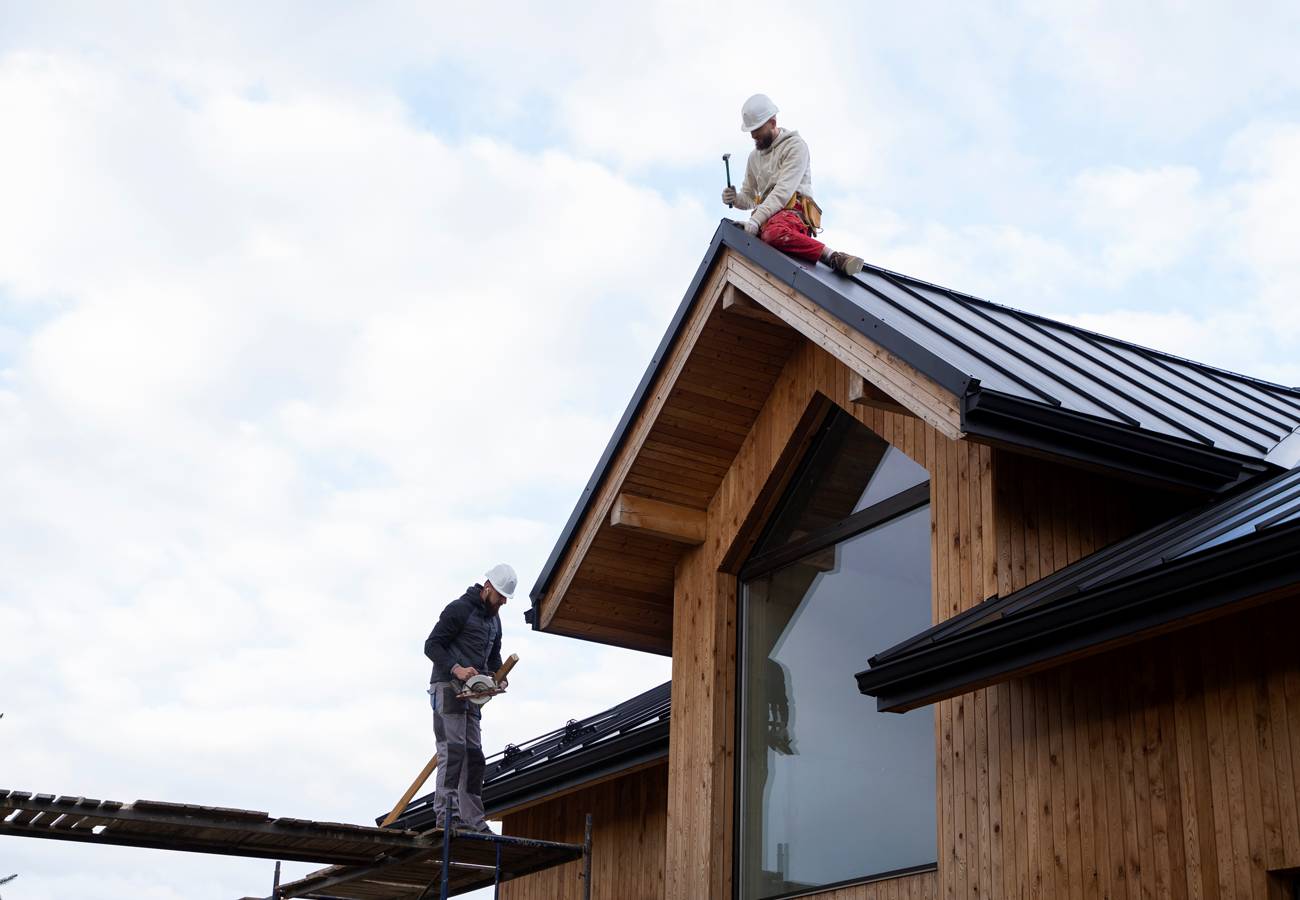 Deux ouvriers travaillent sur la toiture d’une maison en bois, l’un sur l’échafaudage, l’autre au faîtage